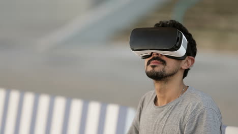 Junger-Arabischer-Mann-Mit-Dunklem-Lockigem-Haar-Und-Bart-Im-Grauen-T-Shirt-Sitzt-Im-Park-Auf-Einer-Gestreiften-Bank-Und-Trägt-Eine-Virtual-Reality-Brille